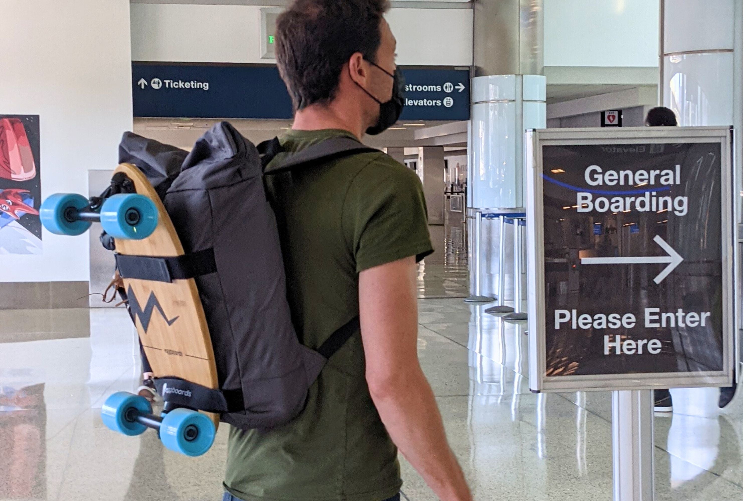 A traveler near a 'General Boarding' sign at the airport, with a mini longboard skateboard strapped to his backpack, showing its portability for travel.
