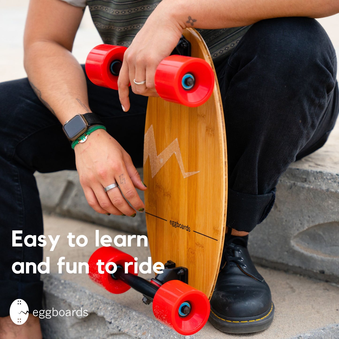 Closeup of a guy sitting on outdoor stairs by the beach, holding an Eggboard mini cruiser skateboard with clear griptape. The image features an overlay stating 'Easy to Learn and Fun to Ride'