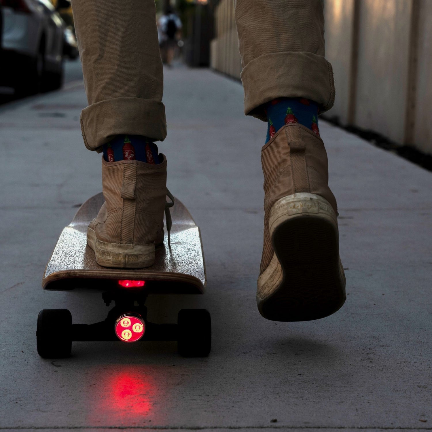 Close-up of a longboard skateboard from behind, featuring a red skateboard light turned on at the back while a guy pushes it forward in an urban landscape. The image highlights the importance of longboard skateboard lights for visibility and safety during night rides.
