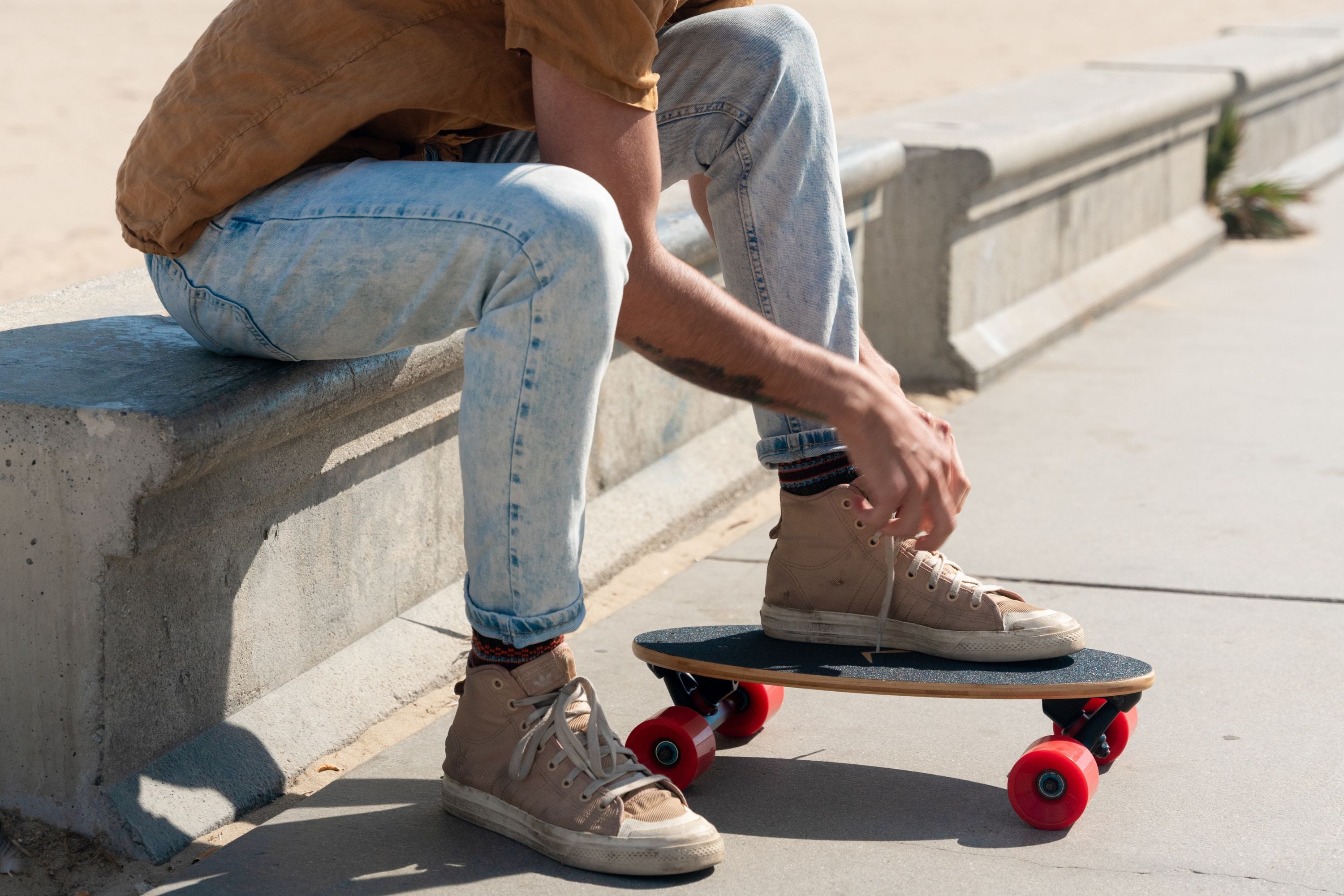 A guy tying his shoes with one foot on a mini longboard skateboard at Venice Boardwalk, capturing a casual beachside skateboarding vibe.