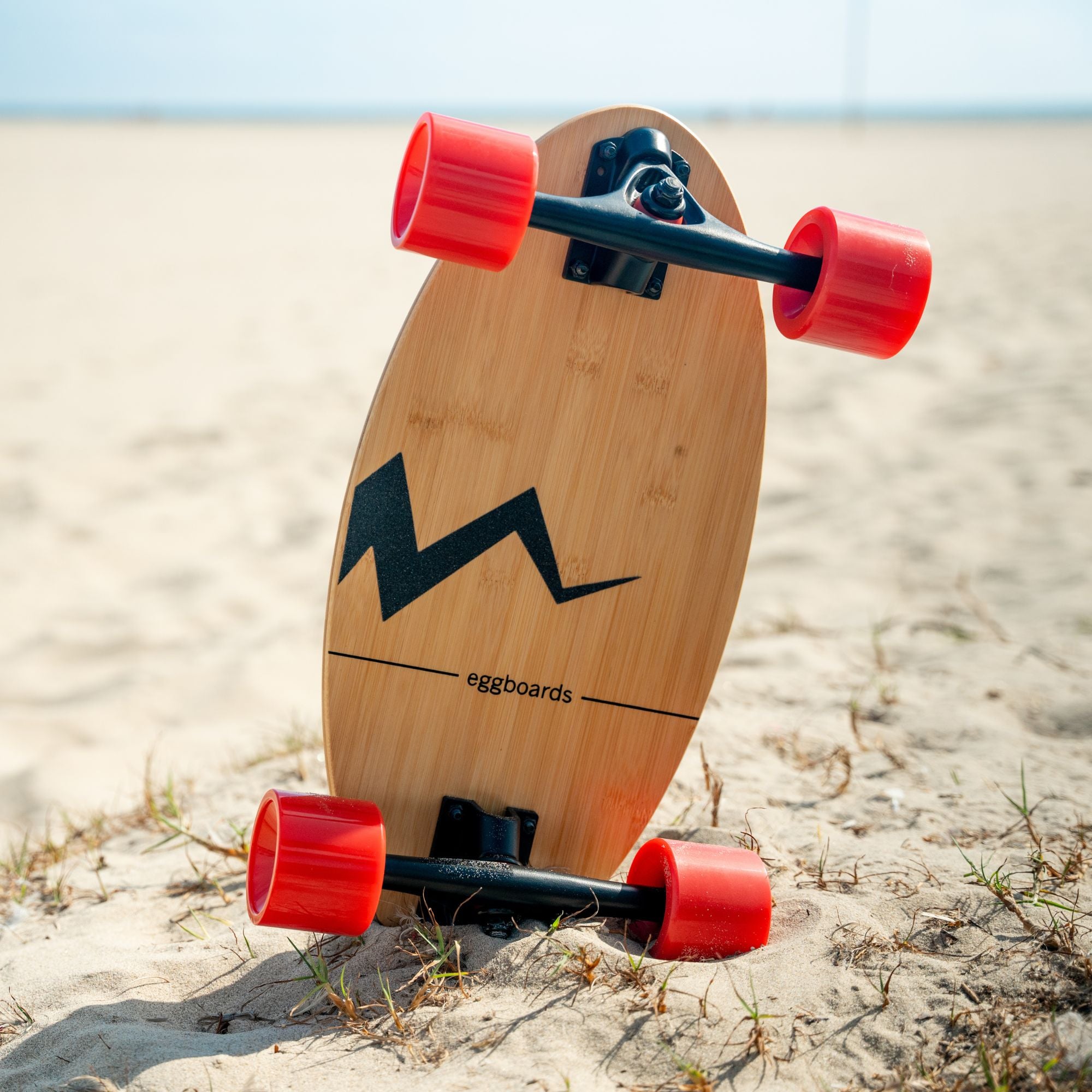 An Eggboard mini longboard skateboard with red wheels resting in the sand by the beach.