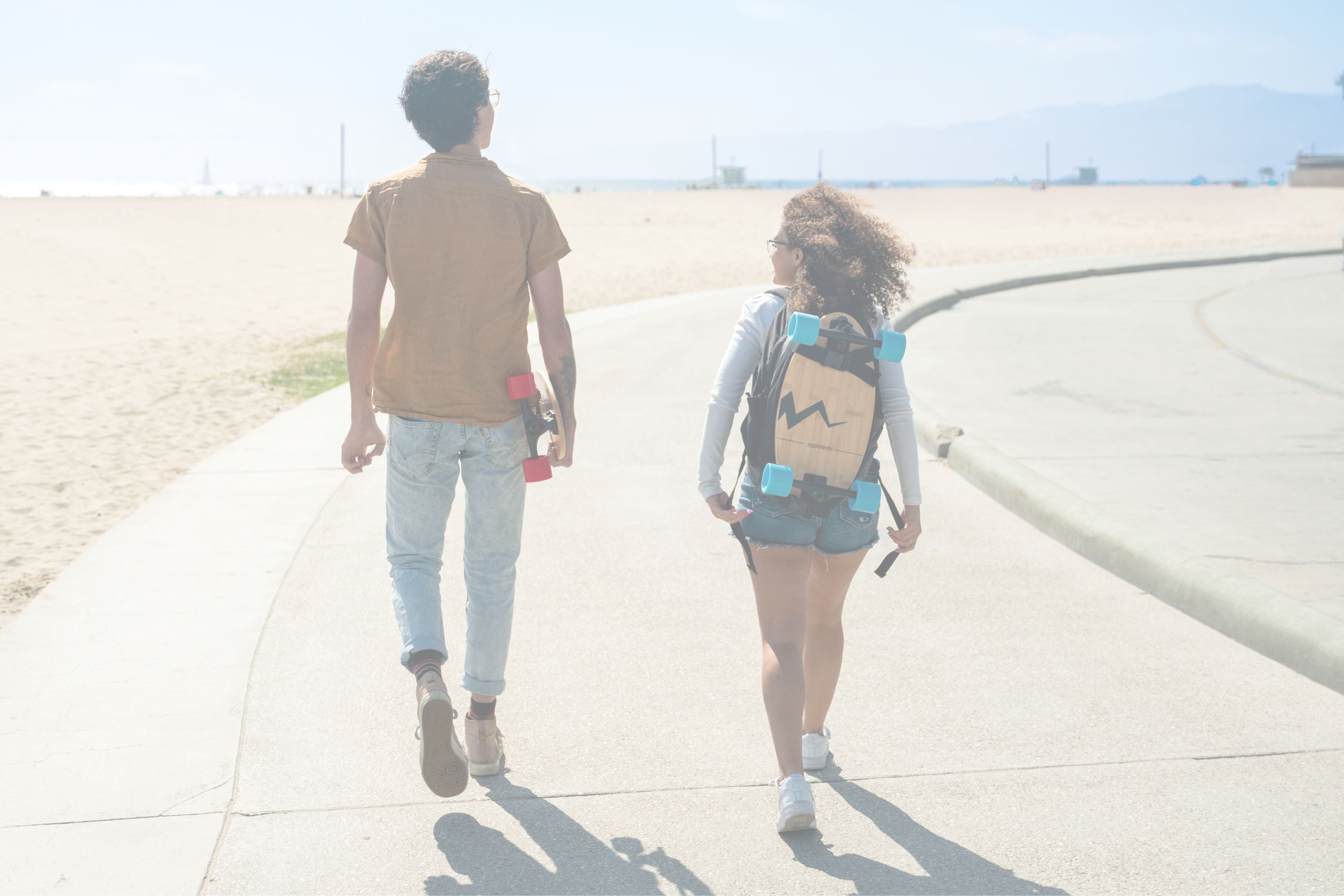 A guy and a girl walking along the Venice Boardwalk, each carrying an eggboard mini longboard skateboard.