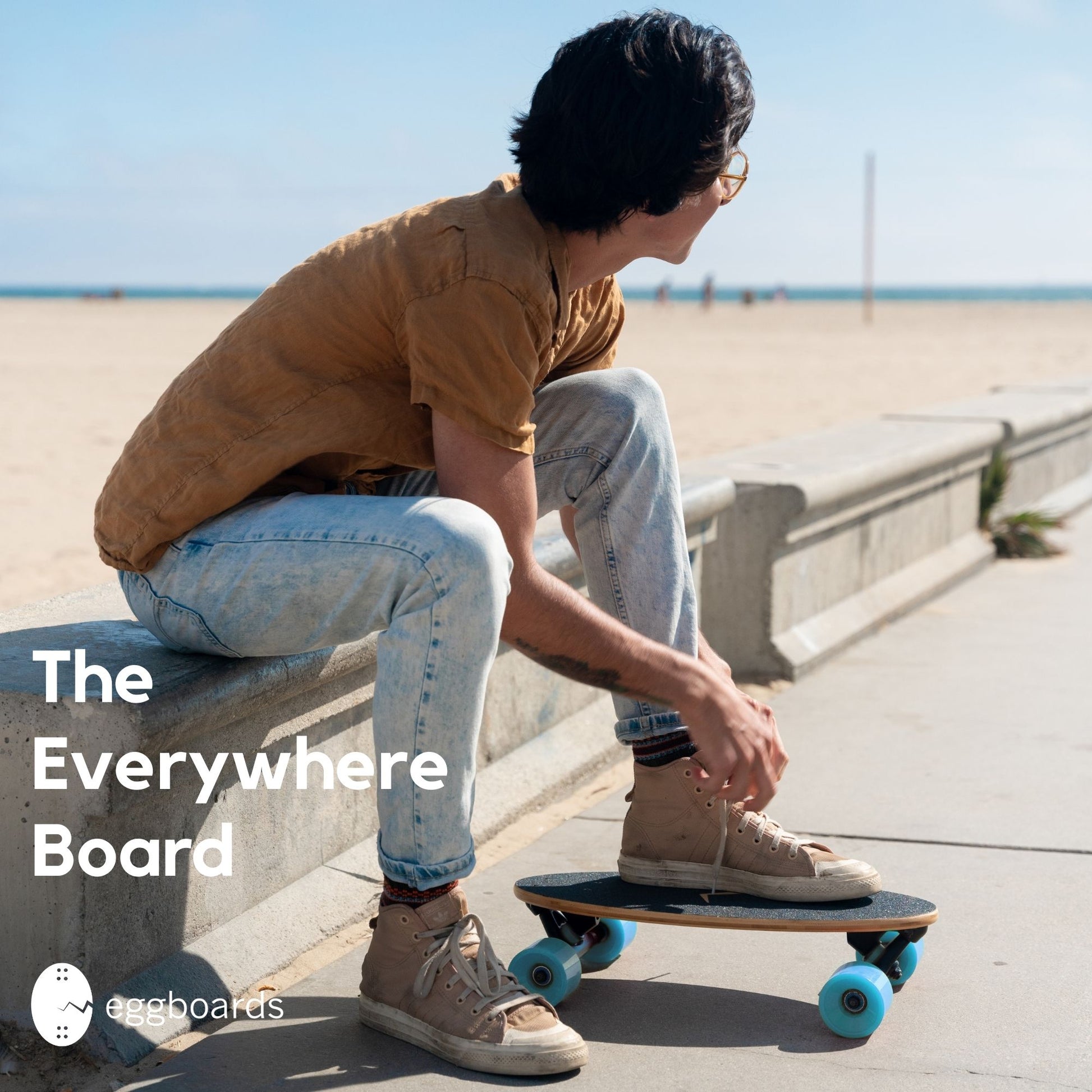 A guy tying his shoes on an Eggboards skateboard cruiser with blue wheels by the beach in Venice, California. The overlay text reads "The everywhere board," emphasizing its versatility and portability for all environments.