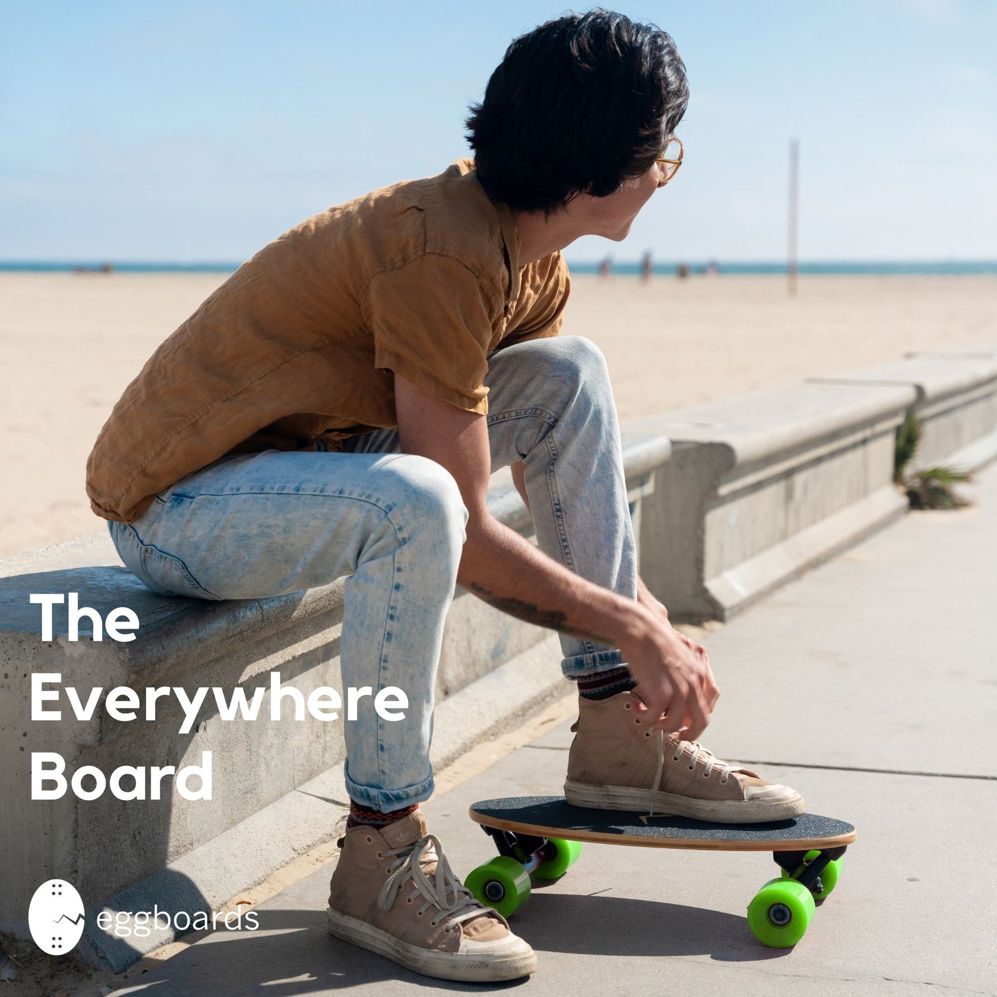 A guy tying his shoes on an Eggboard skateboard cruiser with green wheels by the beach in Venice, California. The overlay text says "the everywhere board."