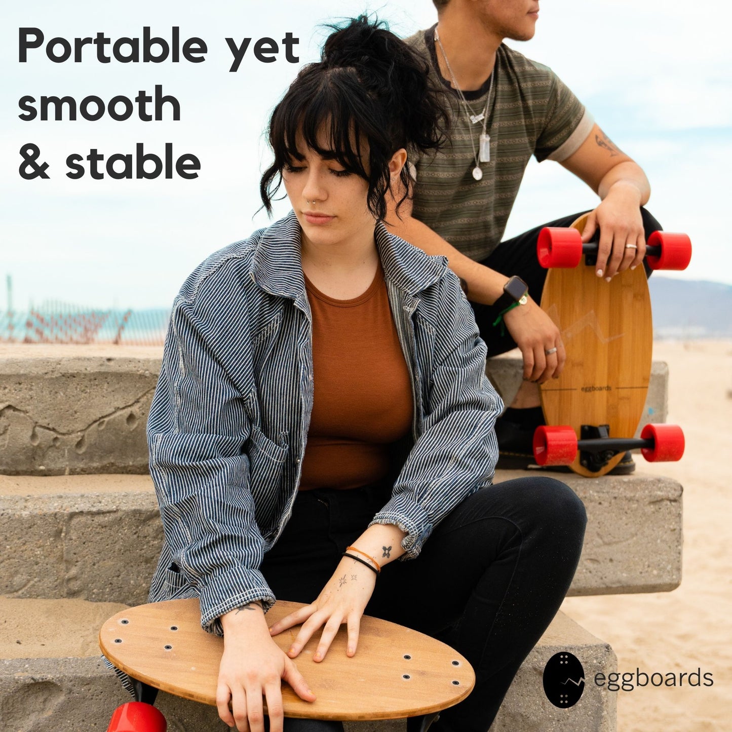 A girl and a guy sitting on stone steps by the beach in Santa Monica, California, with their Eggboard mini longboards resting beside them. The scene captures the relaxed vibe of beach life.