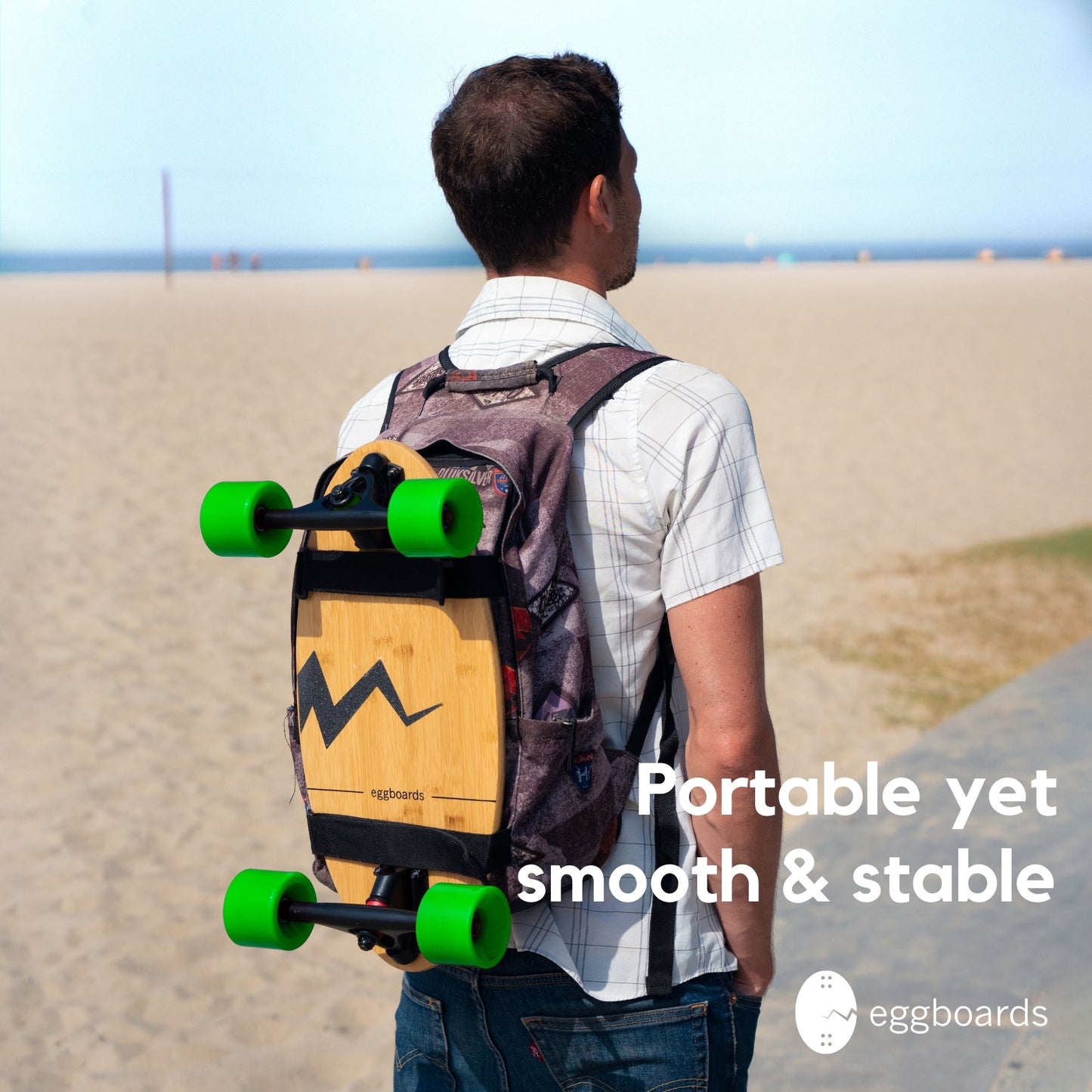 Guy with a skateboard backpack at the beach on a sunny day, carrying an Eggboard mini cruiser with green wheels strapped to his bag. The overlay text says "portable yet smooth and stable.