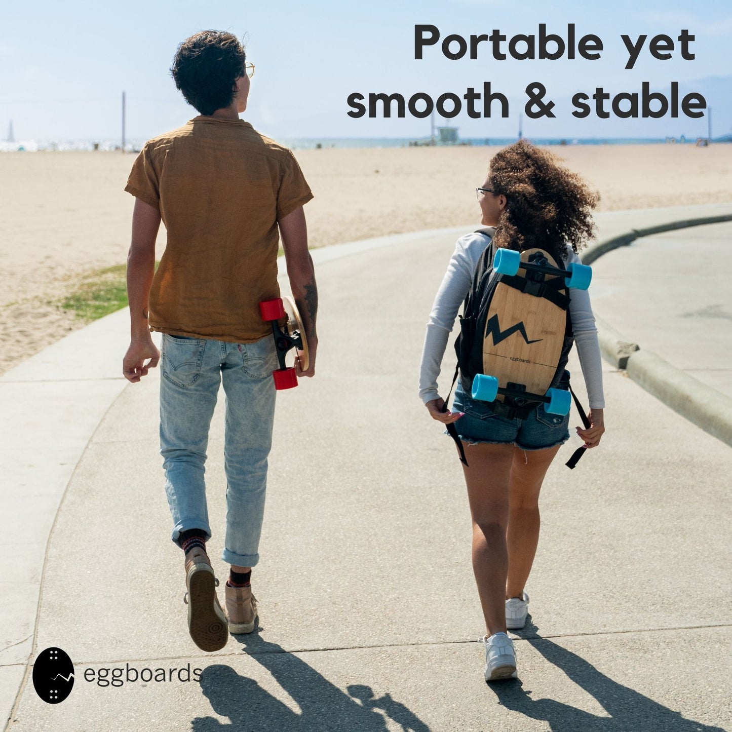 A couple walking by the beach on a sunny day, carrying their Eggboards cruiser skateboards. The guy holds his board by hand, while the girl has hers strapped to her backpack. The overlay text reads "Portable yet stable," highlighting the board's compact design and sturdy build.