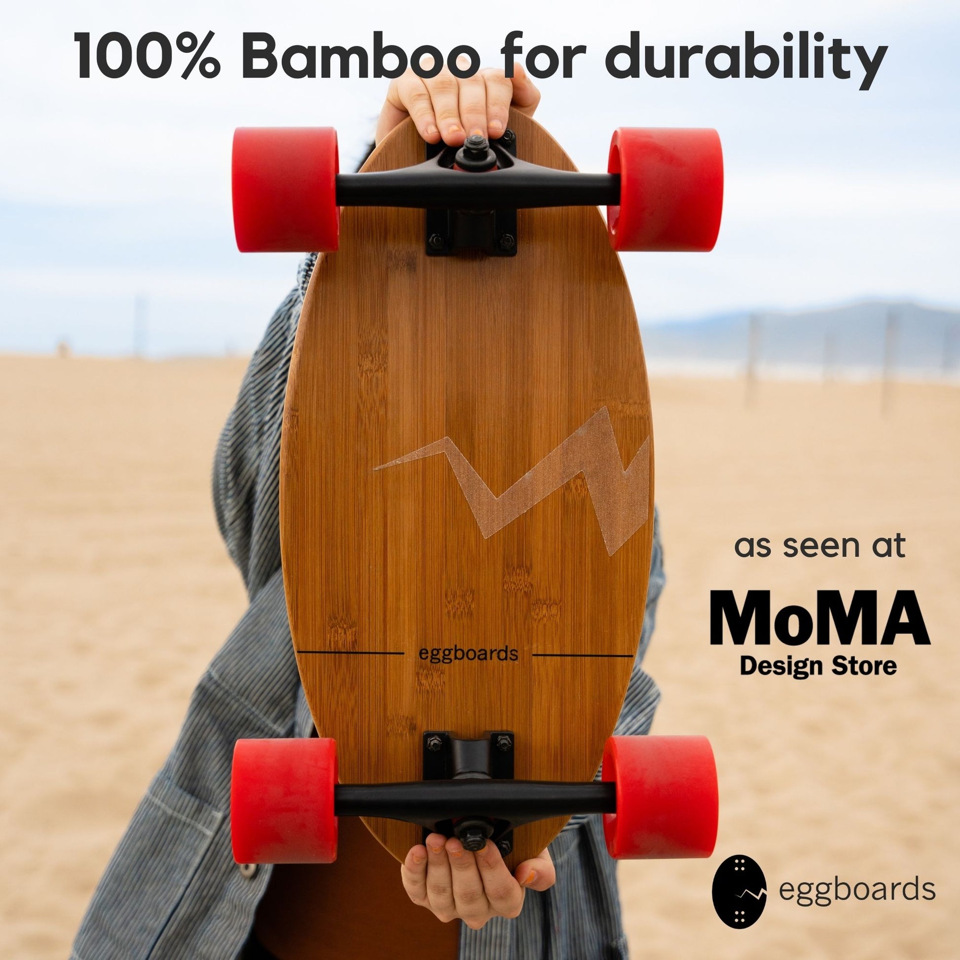 A closeup of a person holding an Eggboard mini cruiser skateboard facing the camera. The board features a bamboo deck showcasing its natural wood grain, red wheels, and clear griptape. The beach in Santa Monica, California, serves as the background, with overlays stating "100% bamboo" and "As seen at the MoMA Design Store.