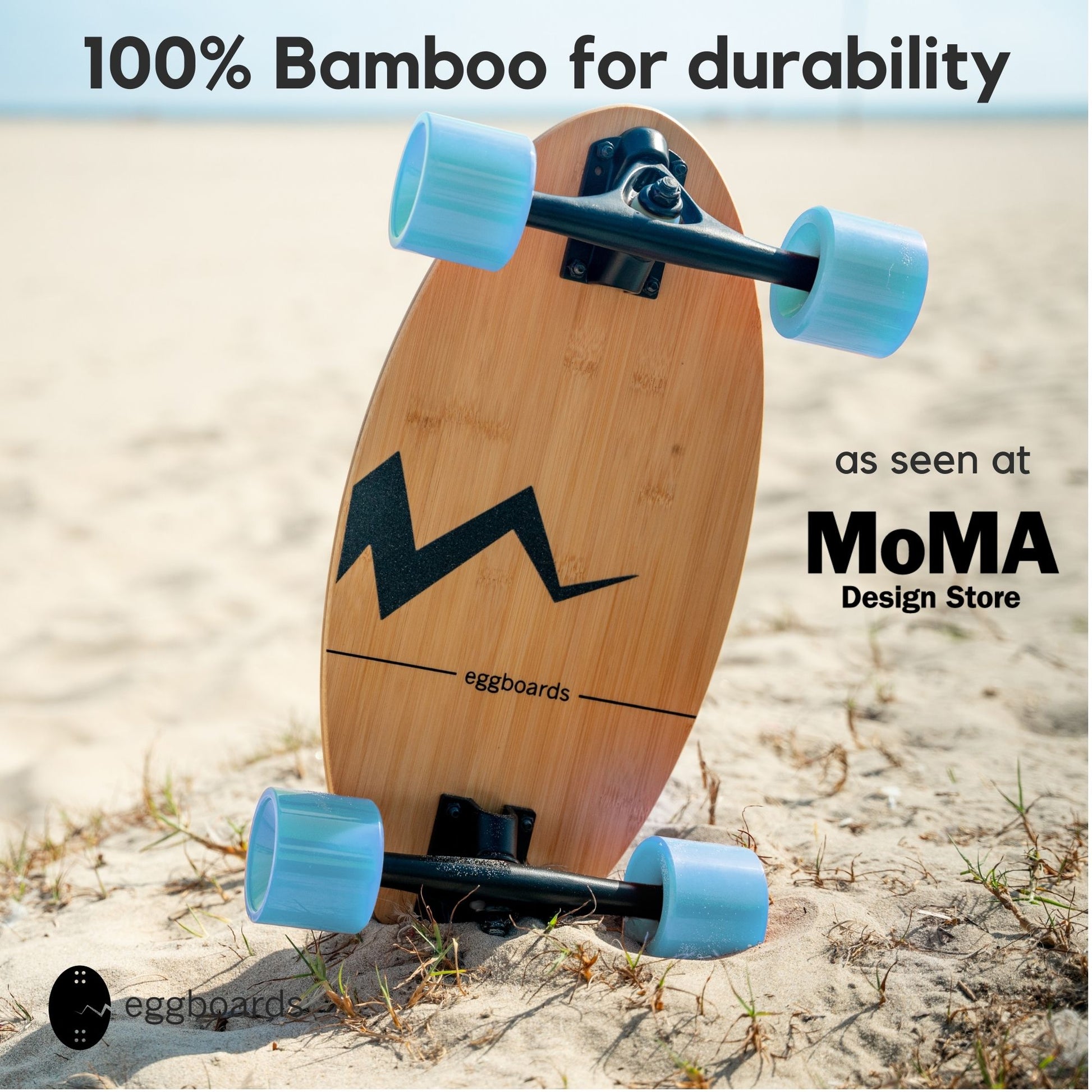 Closeup of the Eggboard bamboo skateboard with blue wheels and a distinctive crack-shaped griptape, resting on the sand at the beach in Venice, California. The overlay text highlights "100% bamboo" and "As seen at the MoMa Design Store," emphasizing the board's premium design and materials